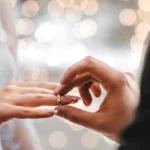 Close-up of a couple exchanging a ring, a hand placing a diamond ring on the partner's finger with soft bokeh lights in the background