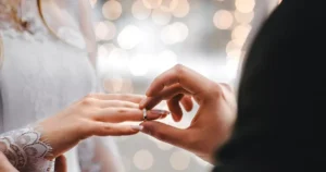 Close-up of a couple exchanging a ring, a hand placing a diamond ring on the partner's finger with soft bokeh lights in the background