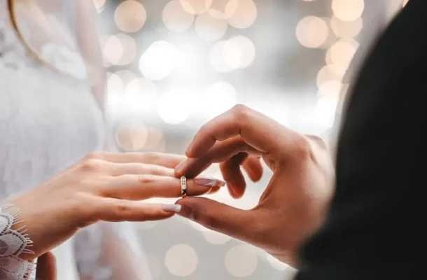 Close-up of a couple exchanging a ring, a hand placing a diamond ring on the partner's finger with soft bokeh lights in the background