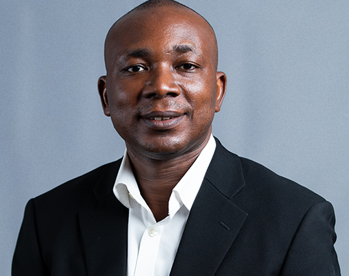 no_translation_8618_e0cy2fufqun7bgm01pwyz9lm_no_translation_translation | Ghanaguardian.com Professional headshot of a smiling man in a black suit and white shirt against a blue backdrop.