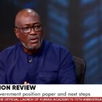Man in a blue shirt wearing glasses speaks in a TV studio about constitution review, with a microphone clipped to his shirt and Joy News branding visible.