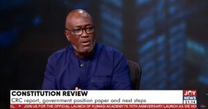 Man in a blue shirt wearing glasses speaks in a TV studio about constitution review, with a microphone clipped to his shirt and Joy News branding visible.