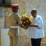 Uniformed officer presents a bright bouquet to a man in white traditional attire at a formal ceremony near a stone wall.