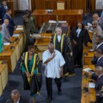 Parliament chamber scene: a man in a white tunic walks down the aisle waving, flanked by officials in ceremonial black robes with gold trim and other lawmakers.