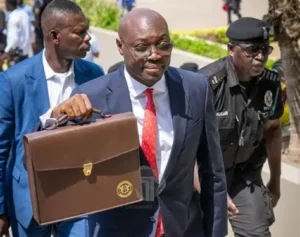 A man in a navy suit with a red tie carrying a brown briefcase, flanked by security guards outdoors.