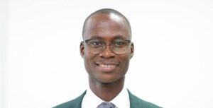 Professional headshot of a smiling man wearing glasses, a white shirt, tie, and green blazer against a white background