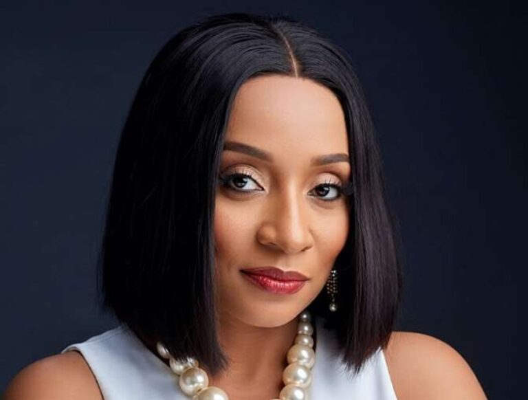 Portrait of a woman with a sleek black bob, wearing a pearl necklace and subtle makeup against a dark background.